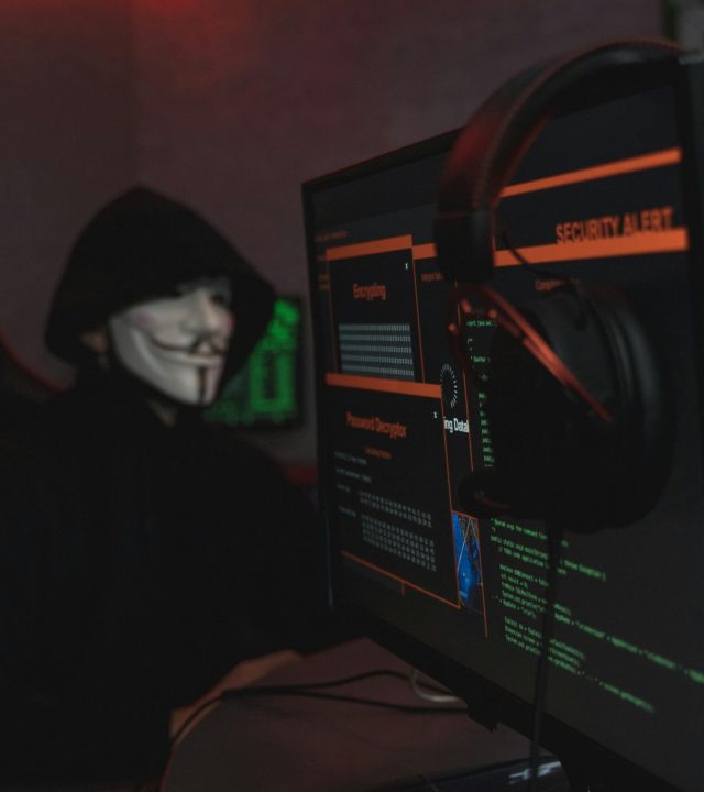 pexels-photo-5380651-5380651 A person wearing a Guy Fawkes mask engaged in hacking activities on a computer in a dimly lit room.